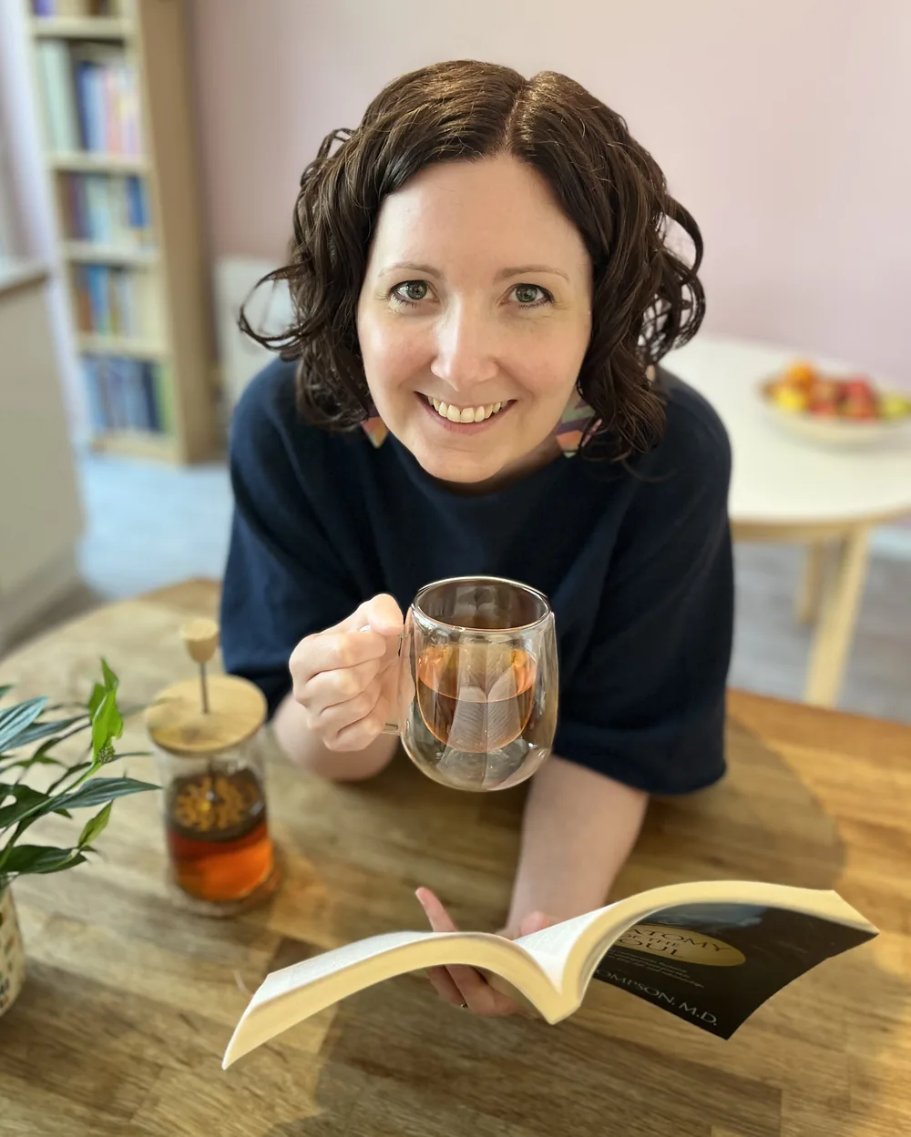 Anna Hobbs at a wooden table with a cup of tea and an open book, smiling warmly